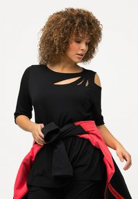 Zwarte lange mouwen top met uitsnijdingen op de schouders en de halslijn. Gecombineerd met een rode en zwarte sweatshirt die aan de taille is geknoopt.