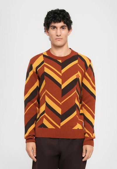 Homme aux cheveux bouclés noirs portant un pull à motifs chevrons rouille, noir et jaune, et un pantalon sombre, se tenant contre un fond uni.