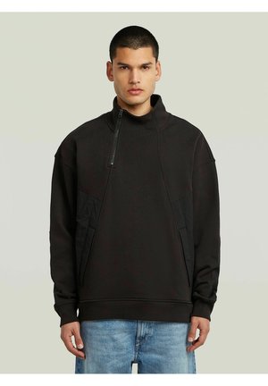 Homme portant un sweat-shirt noir à demi-fermeture éclair avec des poches et un jean bleu, debout devant un fond clair uni.