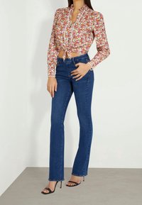 Bloemige blouse met lange mouwen en een strikdetail aan de voorkant, met een mix van rode, roze en gele bloemen. Gecombineerd met blauwe klokvormige jeans en zwarte hakken.