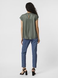 Blouse à manches courtes, olive avec un dos en forme de trou de serrure et des détails froncés, associée à un jean bleu et des sandales à talons carrés noires.