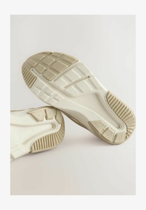 Zapatillas deportivas blancas con suelas texturizadas y suelas de goma con patrón. Incluyen un diseño curvado y acentos en color beige claro.