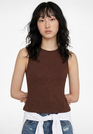 Jeune femme aux cheveux noirs ondulés porte un haut marron sans manches et un jean en denim avec une chemise blanche nouée autour de sa taille.