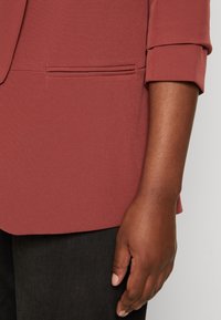 Rostfärgad blazer i glatt tyg med en bröstficka och uppvikta ärmslut, ihopkopplad med mörkgrå byxor.