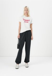 Balta medvilnė marškinėliai su raudonu "TOMMY JEANS" spaudiniu, derinami su juodais sportiniais šortais ir šviesiais slip-on bateliais; elegantiškas dizainas, laisvas siluetas.