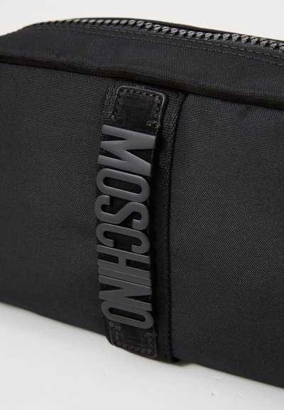 Čierna látková taška s textúrovaným povrchom, na boku s výrazným čiernym gumovým logom "Moschino" a so zipsom na vrchu.