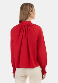 Rote Bluse mit hohem Kragen, die über eine geraffte Rückendetails und lange, smockierte Ärmel verfügt. Glattes Material mit lockerer Passform.