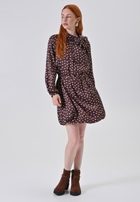 Braunes Kleid mit Schleifenband am Ausschnitt und weißen Polka-Dots. Lange Ärmel, tailliert und ausgestellte Silhouette. Braune Stiefeletten.