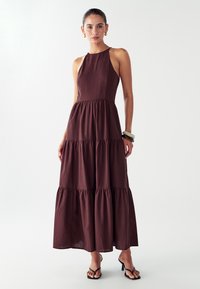 Nainen, joka on pukeutunut hihattomaan, kerrokselliseen tummanruskeaan maksimekkoon, jossa on kultaiset rengaskorvakorut, kerroksittain käytettyjä rannekoruja ja mustat korkokengät, valkoista taustaa vasten.