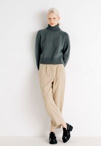 Dark green knit turtleneck sweater with ribbed hem, paired with beige pleated trousers and black chunky soled shoes.