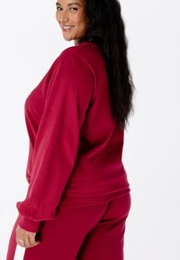 Femme souriante avec de longs cheveux noirs portant un sweat-shirt et un pantalon rouges assortis, légèrement tournée sur le côté devant un fond blanc.