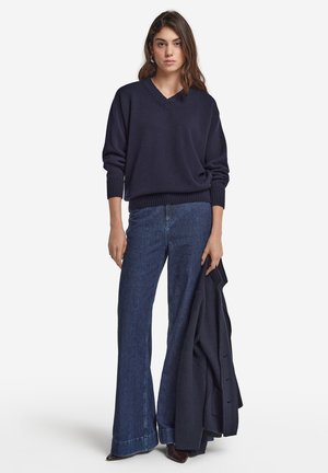 Dunkelblauer V-Ausschnitt-Pullover mit gerippten Bündchen, kombiniert mit hochgeschnittenen blauen Schlaghosen. Das Modell hält eine passende dunkelblaue Jacke.