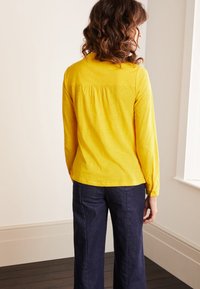 Top amarillo de manga larga con detalle fruncido en la parte posterior, combinado con pantalones de pierna ancha en azul oscuro. La tela parece suave y con un ligero brillo.