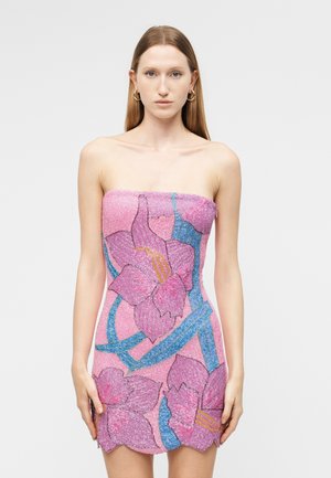 Kobieta w różowej mini sukience na ramiączkach, ozdobionej dużymi niebiesko-fioletowymi wzorami kwiatowymi, stojąca na tle jednolitej białej ściany.