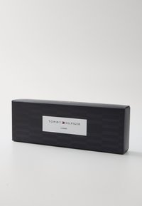 Caja rectangular negra con un patrón texturizado, que presenta una etiqueta blanca que dice "TOMMY HILFIGER 3 PARES". Diseño simple, líneas limpias.
