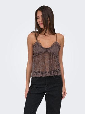 Vrouw draagt een bruine doorschijnende kanten camisole met dunne bandjes en zwarte hoge taille broek, poserend tegen een effen lichte achtergrond.