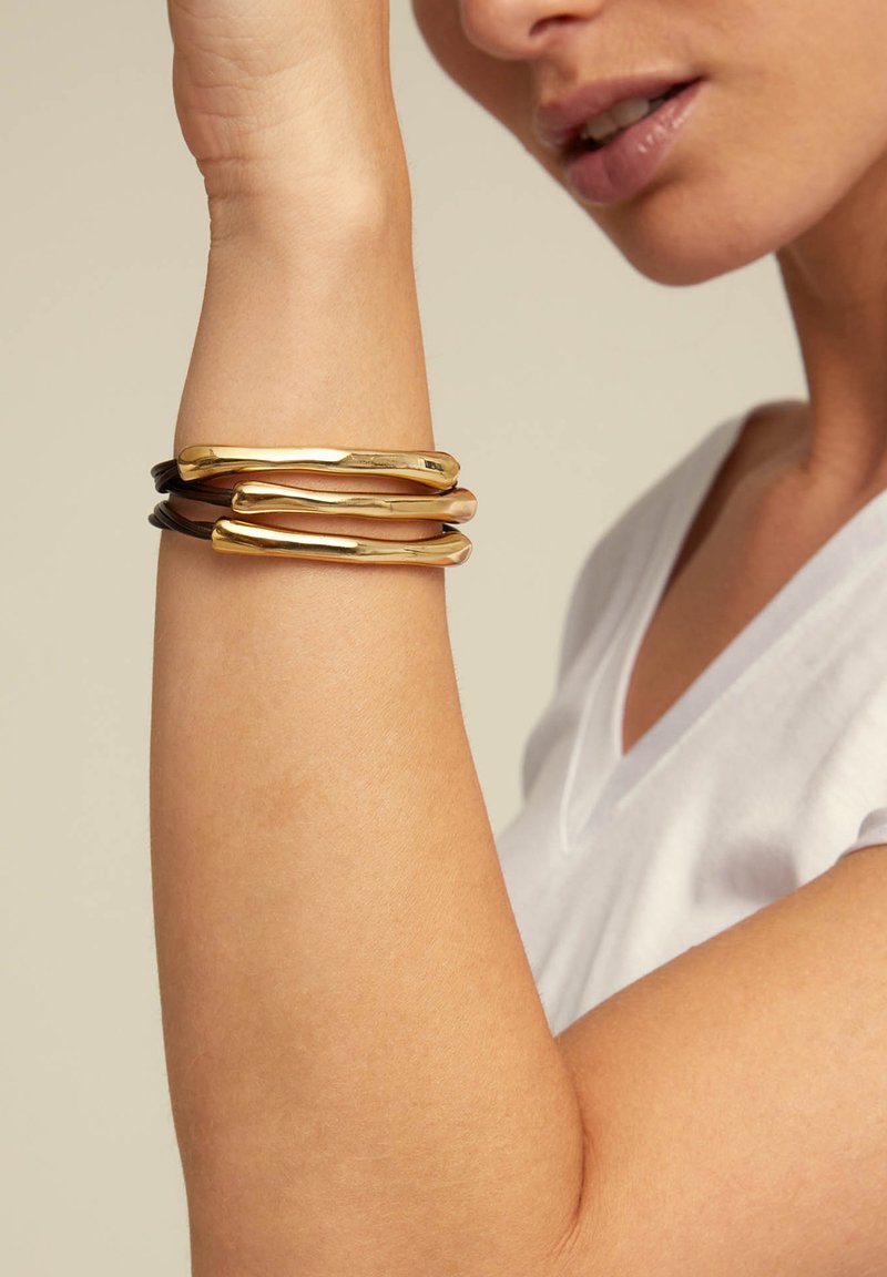 Des bracelets en or et en bronze sur un poignet, avec des épaisseurs et des formes variées. Finition en métal lisse avec un design légèrement ondulé. Chemise blanche décontractée.