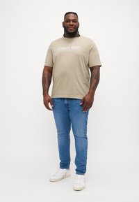 Bež majica od pamuka s logotipom "Calvin Klein", u kombinaciji s plavim trapericama i bijelim tenisicama. Model stoji nasuprot neutralnoj pozadini.