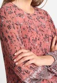 Blusa de manga larga con estampado de rosas, textura plisada y puños elásticos. Presenta un escote redondo y un intrincado diseño floral en toda la prenda.