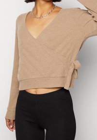 Beige ribbat stickad omslagstopp med långa ärmar, djup V-ringning och sidoknytdetalj, i kombination med svarta leggings. Texturerad och figurnära design.