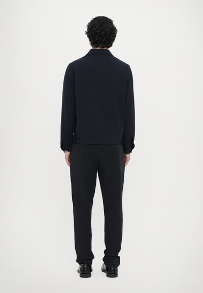 Moška temno modra jakna, krojena prileganja, z ovratnikom in gumbi na hrbtni strani, v kombinaciji z črnimi hlačami in čevlji. Gladka tekstura.