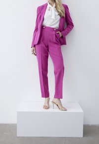Costume sur mesure fuchsia avec un blazer et un pantalon taille haute, associé à un chemisier blanc et des talons dorés à lanières, posé sur un piédestal blanc.