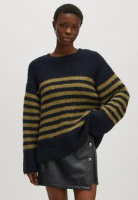 Navy- und olivgestreifter Strickpullover mit lockerer Passform, weiten Ärmeln und gerippter Textur, kombiniert mit einem schwarzen Minirock aus Kunstleder mit silbernen Knöpfen.