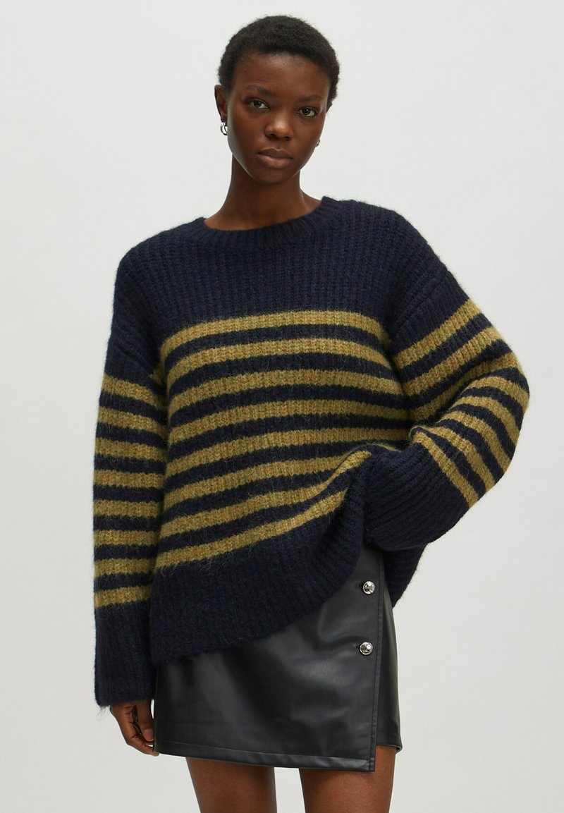 Navy- und olivgestreifter Strickpullover mit lockerer Passform, weiten Ärmeln und gerippter Textur, kombiniert mit einem schwarzen Minirock aus Kunstleder mit silbernen Knöpfen.