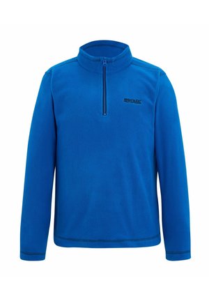 Ragazzo in piedi che indossa un pile blu con zip, pantaloni cargo neri e scarpe sportive nere e verde neon su uno sfondo chiaro e semplice.