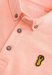 Camisa polo texturizada rosa con cuello, que presenta botones oscuros y un bordado de langosta amarilla en el lado izquierdo del pecho. Nombre de la marca en el interior del cuello.
