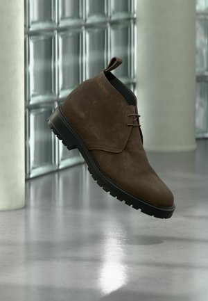 Stivaletto alla caviglia in camoscio marrone con suola nera e lacci che fluttua sopra un pavimento riflettente vicino a una parete in blocchi di vetro e un pilastro bianco.