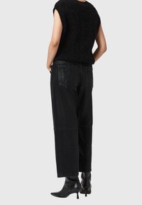 Personne portant un top noir texturé sans manches, un pantalon noir large avec poches plaquées, et des bottines noires à talons, debout sur un fond blanc.