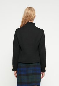 Μαύρο εφαρμοστό σακάκι με ψηλό γιακά και δομημένους ώμους, με διακοσμητικά χρυσά κουμπιά στο μανίκι. Φορεμένο πάνω από καρό φούστα.