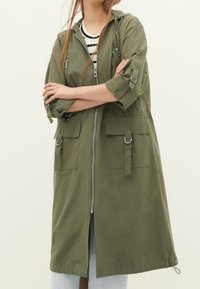 Olive green lightweight parka jacket with a front zipper, adjustable hood, and two flap pockets. Features roll-up sleeves and drawstring hem.