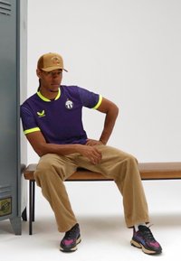 Lila sportströja med korta ärmar och neon-gula accenter, kombinerad med beige manchestertygbyxor och flerfärgade sneakers. Sitter på en bänk.