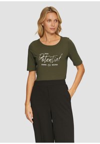 Olijfgroene t-shirt met korte mouwen met een zilverkleurige metallic print met de tekst "JOUW potentieel KENT geen BRENGEN." Gecombineerd met zwarte high-waisted broek.