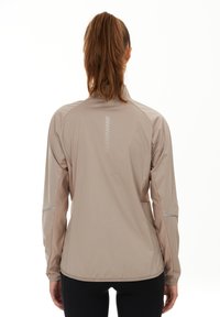 Ljust beige jacka med långa ärmar, mjuk textur, reflexdetaljer och hög krage. Baksidan har vertikala linjedetaljer för ökad synlighet.