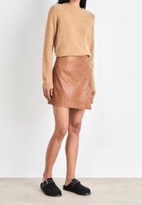 Pull en tricot beige à manches longues, porté sur une mini-jupe en cuir marron fermée par une fermeture éclair latérale ; mules en daim noir avec des clous décoratifs.