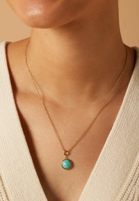Collier en métal doré avec un pendentif rond en turquoise. Le pendentif a une surface lisse et est monté dans un cadre simple. La chaîne est délicate et fine.