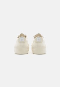 Sneakers in tela beige con suola piatta, caratterizzati da un logo a stella bianca sul tallone e dettagli testurizzati. Vista posteriore del design.
