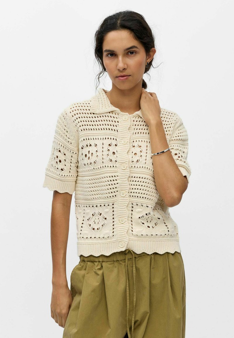Femme aux cheveux foncés porte un haut crème à manches courtes en crochet avec boutons et un pantalon ample vert olive, debout devant un fond uni.