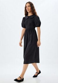 Robe noir, longueur genou, avec un col rond, des manches bouffantes et une taille froncée. Le tissu semble lisse avec des détails de couture verticaux.
