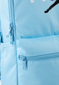 Light blue backpack made of textured fabric features multiple zipped pockets, a padded section, and a logo in contrasting dark color.