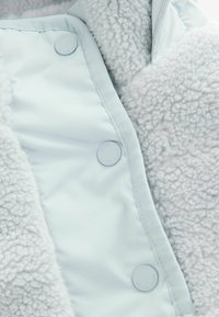 Ljusblå jacka med mjuk, texturerad fleece på ena sidan och slät tyg på den andra, fastsatt med runda tryckknappar.