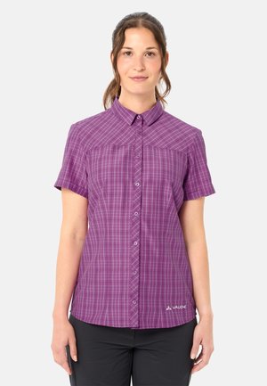 Frau trägt ein kurzärmeliges lila kariertes Hemd mit Knöpfen und schwarze Hose, steht vor einem schlichten hellen Hintergrund.