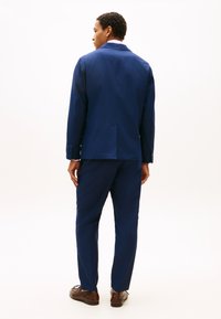 Άνδρας με μπλε σκούρο κοστούμι και καφέ παπούτσια, στέκεται γυρισμένος με την πλάτη προς την κάμερα σε ένα λευκό φόντο.