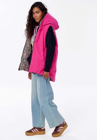 Růžový kordový vest s kapucí, zdobený podšívkou s leopardím vzorem. V kombinaci s černým mikinou, světlými modrými širokými džíny a hnědými teniskami.