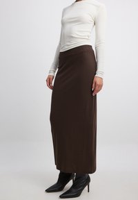 Jupe longue marron avec une taille ajustée et un matériau doux et texturé. Associée à un haut à manches longues léger et des escarpins noirs à bout pointu.