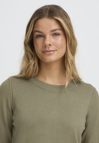 Olivzaļa adīta džempera ar apaļu kakla izgriezumu, garām piedurknēm un teksturētu audumu. Vienkāršs dizains bez redzamām rakstu vai akcentu detaļām.