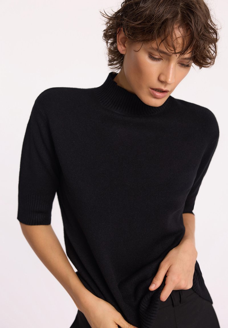 Czarny sweter dzianinowy z wysokim ściągaczem i krótkimi rękawami, o luźnym kroju i miękkiej fakturze. Ograniczone widoczne detale.